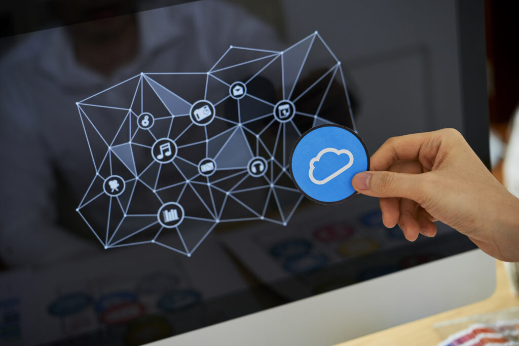 Hand holding a cloud icon in front of a computer screen displaying a network of interconnected digital services, representing the concept of Cloud Projects for storing, sharing, and managing data online.